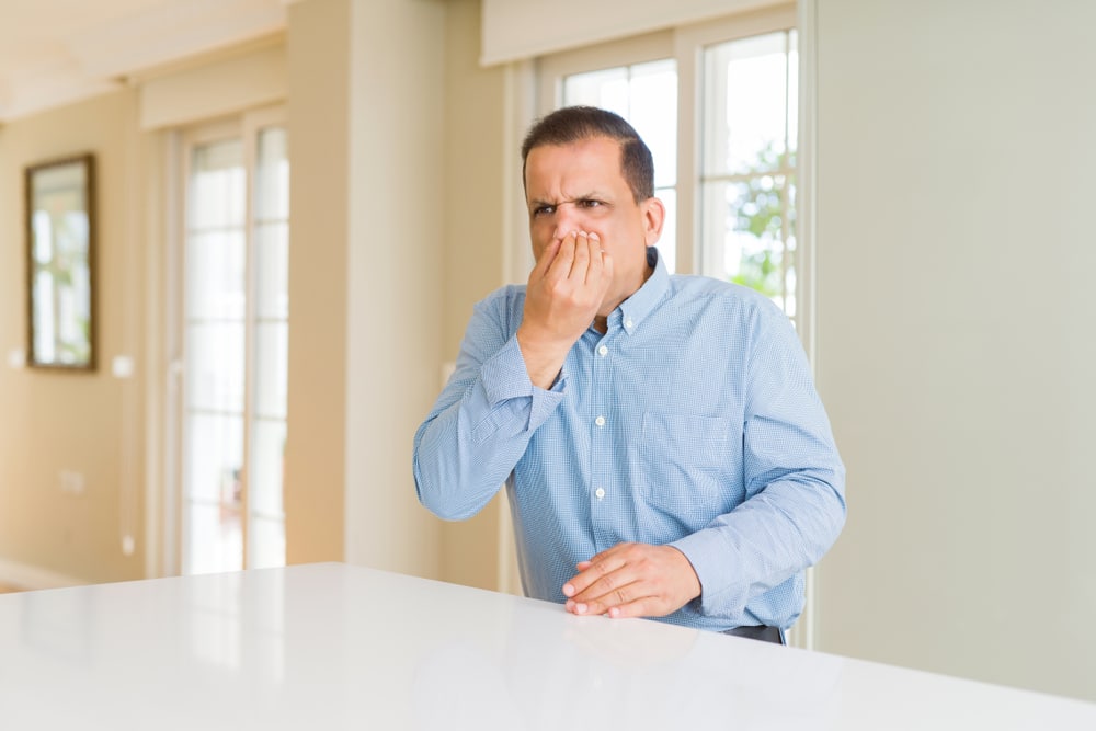 A man in a blue button-up shirt sits at a white table indoors, holding his nose with a displeased expression, as if reacting to a sewer gas smell. Large windows and light-colored walls are visible in the background. | High 5 Plumbing, Heating & Cooling