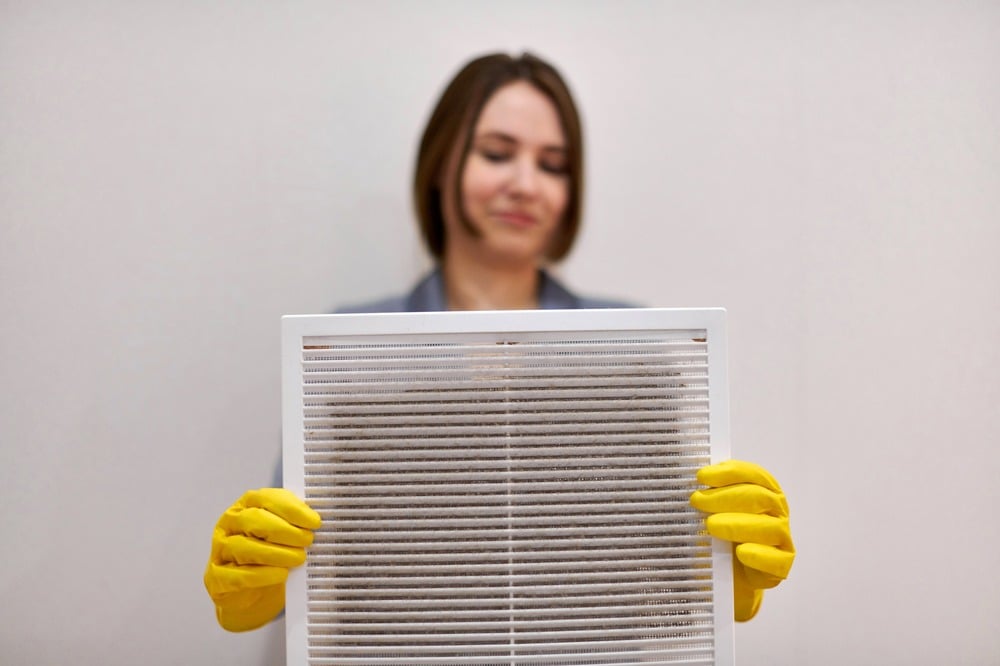 A person wearing yellow cleaning gloves holds up a dirty air filter, showing dust and debris that impact air filter efficiency. The background is plain and out of focus. | High 5 Plumbing, Heating & Cooling
