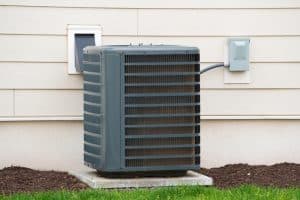 A central air conditioning unit sits on a concrete pad outside a house, with electrical wiring connected to a nearby box on the exterior wall. AC units are known for using the most electricity, while the ground is covered with mulch and grass. | High 5 Plumbing, Heating & Cooling