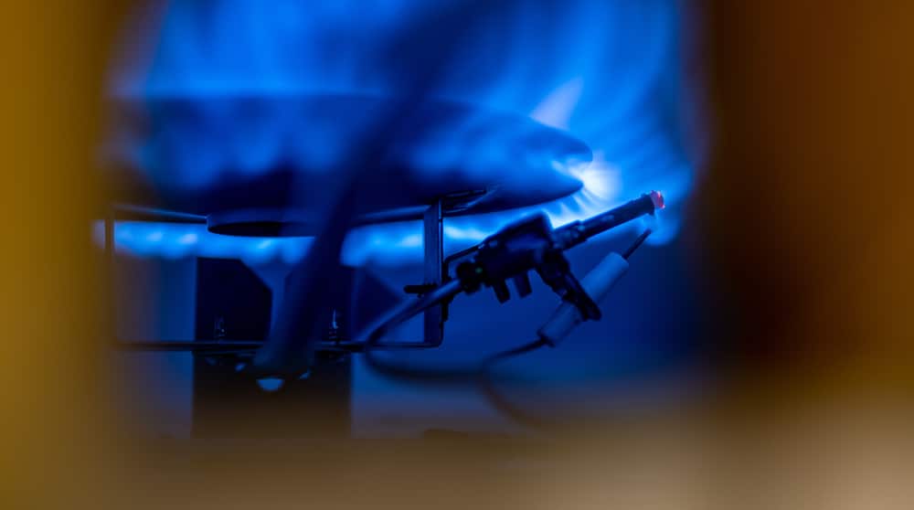 A close-up view of a lit blue gas flame on a stovetop burner, with an igniter and wires visible in the foreground, highlights the pilot light importance for safely igniting and maintaining the burner flame. | High 5 Plumbing, Heating & Cooling