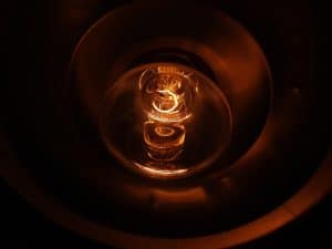 A close-up view of an illuminated incandescent light bulb, glowing orange inside a metallic socket, with the filament clearly visible—a scene often seen when investigating reasons for flickering lights or other home electrical issues. | High 5 Plumbing, Heating & Cooling