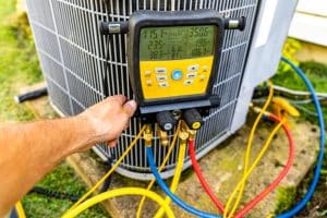 A person’s hand holds a digital manifold gauge attached to an outdoor HVAC unit with yellow, blue, and red hoses for AC troubleshooting. The gauge displays pressure and temperature readings to help diagnose AC common causes or an AC not cooling issue. | High 5 Plumbing, Heating & Cooling