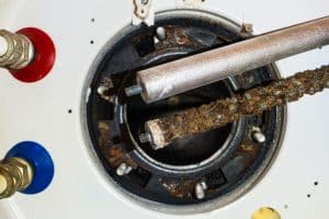 Two water heater anode rods are shown above an open heater tank—one new and clean, the other old and heavily corroded. Visible red and blue pipe connections highlight the importance of regular Water Heater Maintenance and Anode Rod Replacement. | High 5 Plumbing, Heating & Cooling