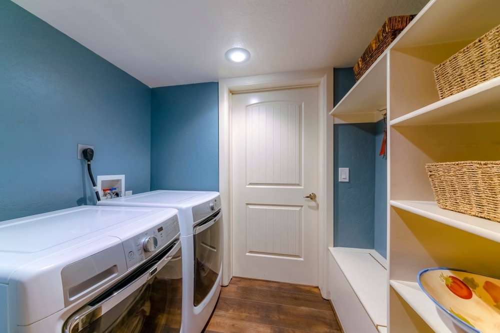 A laundry room with blue walls, a white door, front-loading washer and dryer, white shelving units with woven baskets, a bench, and a decorative bowl on a lower shelf. The wood floor complements the setup, which may use a 3 prong dryer outlet. | High 5 Plumbing, Heating & Cooling