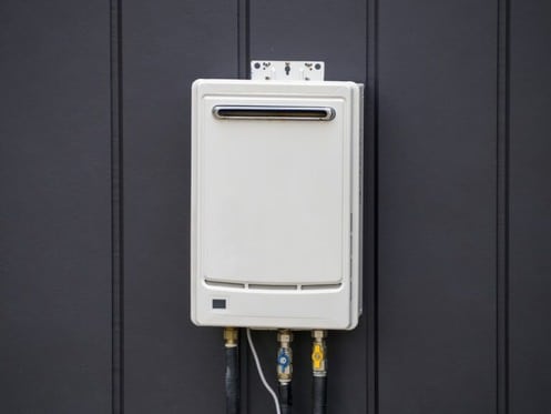 A white tankless water heater is mounted on a dark vertical-panel wall, with several pipes and fittings extending from the bottom—ideal access for essential tankless water heater maintenance such as flushing the tankless heater. | High 5 Plumbing, Heating & Cooling