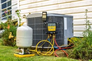 An air conditioning unit sits outside a house, connected to various hoses and diagnostic gauges. A refrigerant tank is nearby on the grass. The unit is next to a white siding wall with some plants visible around it. | High 5 Plumbing, Heating & Cooling