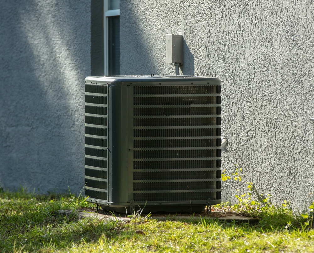 An ideal air conditioning unit with a high SEER rating sits on a concrete slab next to a gray-textured exterior wall, surrounded by grass and small yellow flowers. | High 5 Plumbing, Heating & Cooling