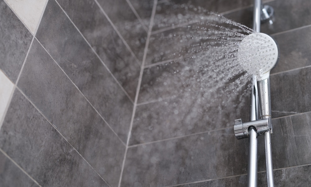 A close-up view of a showerhead spraying water at an angle in a tiled shower with grey and beige tiles on the walls, highlighting shower durability for a longer shower life span. | High 5 Plumbing, Heating & Cooling
