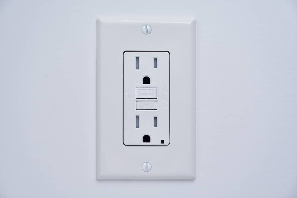 A white electrical outlet with two sockets and two rectangular buttons, mounted on a white wall. This tamper-resistant GFCI outlet offers enhanced safety features to help protect against electrical shocks. | High 5 Plumbing, Heating & Cooling