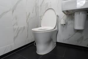 A modern white toilet with the lid open—ideal for toilet troubleshooting like when the toilet tank is not filling—sits next to a wall-mounted sink and toilet paper holder in a bathroom with marble-patterned walls and a black tiled floor. | High 5 Plumbing, Heating & Cooling