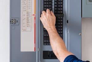 A person’s hand adjusts a circuit breaker switch on an open panel, practicing electrical safety. The panel has labeled switches and a list of circuits attached to the inside of the door, assisting with circuit breaker troubleshooting. | High 5 Plumbing, Heating & Cooling